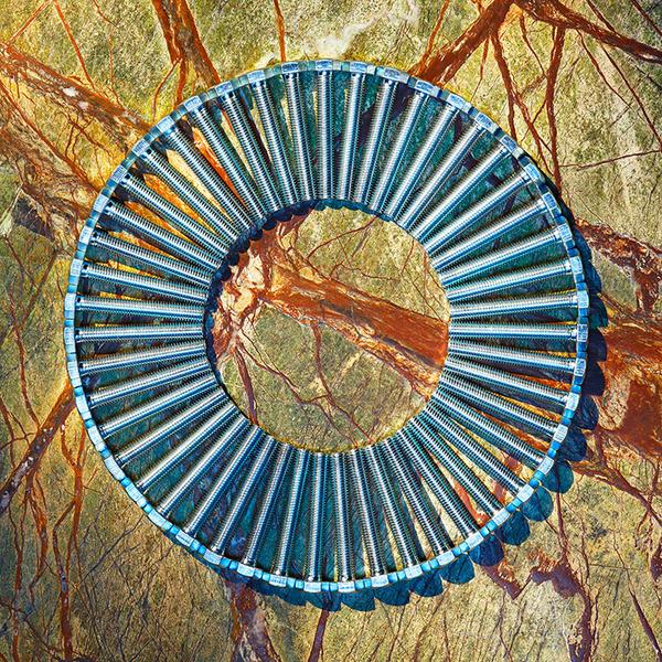 Ein kreisförmiges Objekt, bestehend aus vielen Schrauben, liegt auf einer strukturierten Oberfläche mit erdigen Farben und Linien. Die Anordnung des Objekts schafft einen interessanten Kontrast zur darunterliegenden Textur.