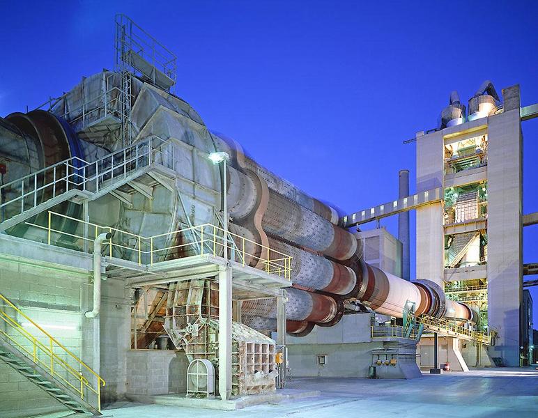 Industrieanlage bei Nacht, mit einem großen rotierenden Kamin, der in einer modernen Betonanlage eingebaut ist. Säulen und Treppenverläufe sind sichtbar, sowie verschiedene Maschinen und Förderanlagen. Die Umgebung ist in kühlem Blau beleuchtet.