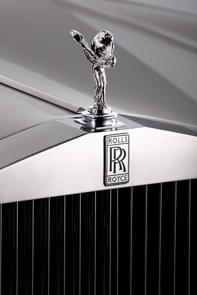 Eine Nahaufnahme des Kühlergrills eines Rolls-Royce mit dem emblematischen "Spirit of Ecstasy" (eine stilisierte Figur) und dem Rolls-Royce-Logo. Der Grill ist glänzend und hat vertikale Streifen.