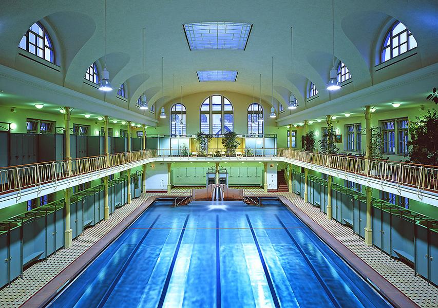 Ein großer, eleganter Innenraum eines Schwimmbads mit einem blauen Schwimmbecken in der Mitte. Die Wände sind mit Fenstern und Pflanzen geschmückt, und große, gewölbte Decken sorgen für eine helle, offene Atmosphäre.