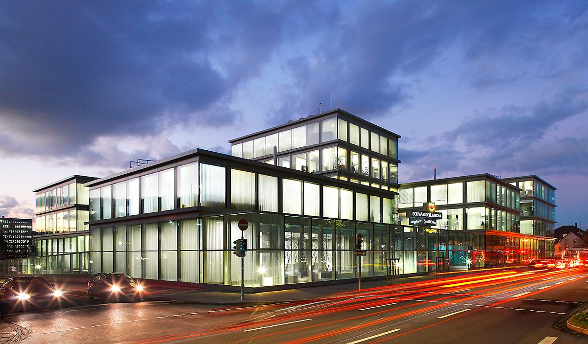 Moderne Bürogebäude mit Glasfassade bei Nacht, beleuchtet von Innen. Straßenverkehr ist sichtbar, mit Lichtspuren von fahrenden Autos. Der Himmel ist bewölkt und es ist Dämmerung.