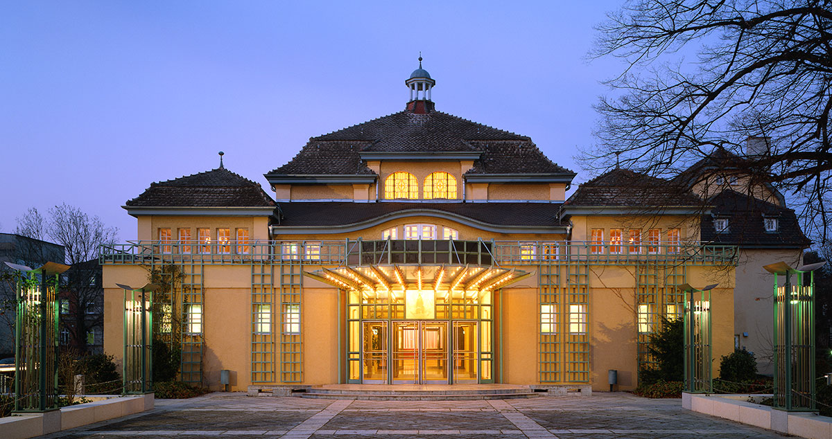 Historisches Gebäude mit stilvollem Eingang, beleuchtet in der Dämmerung. Die Fassade zeigt eine Mischung aus traditioneller und moderner Architektur, umgeben von Bäumen und einem geräumigen Vorplatz.