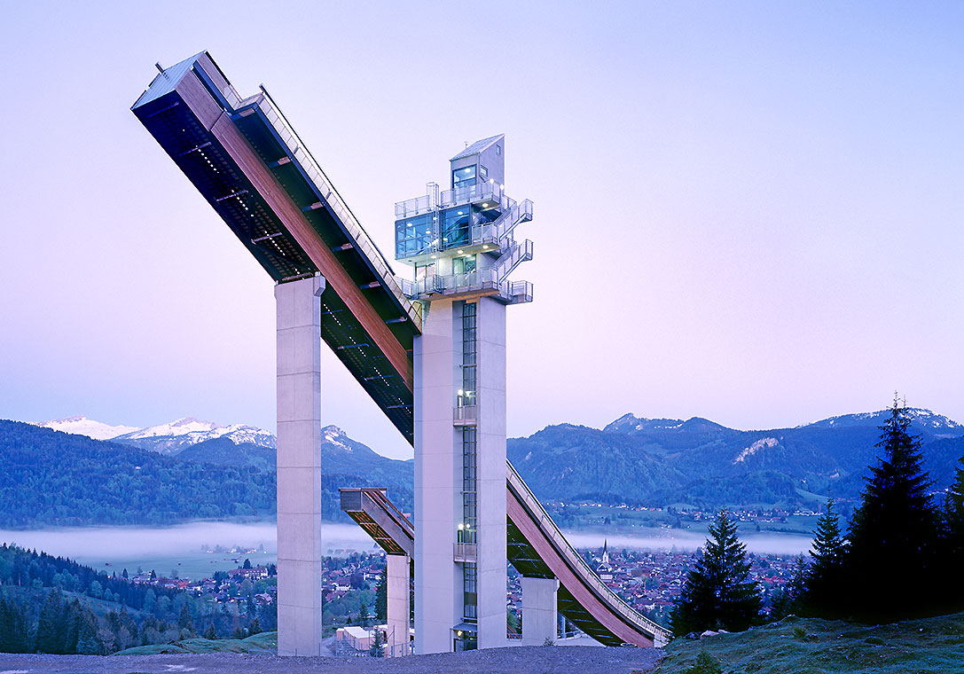 Eine Skisprungschanze steht majestätisch vor einem sanften Bergpanorama. Der hellblaue Himmel im Hintergrund verdeutlicht die Morgenstunde, während die umliegenden Wälder und das Tal leicht in Nebel gehüllt sind.