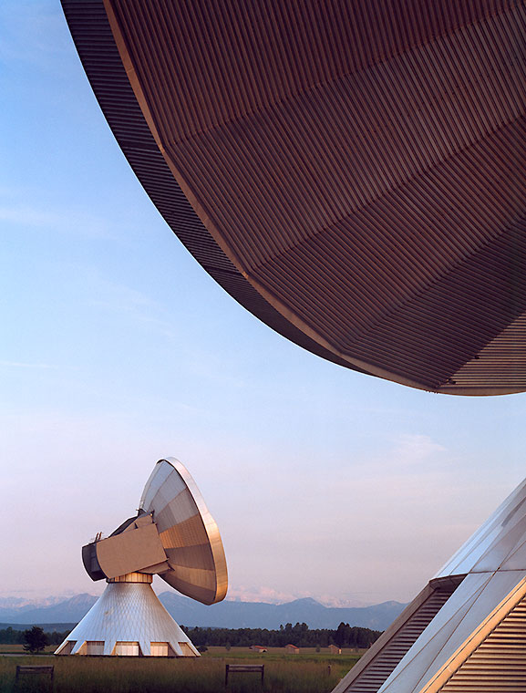 Zwei große Parabolantennen stehen auf einem offenen Feld. Die vordere Antenne ist leicht geneigt, während die hintere Antenne schräg im Hintergrund sichtbar ist. Der Himmel ist klar und zeigt sanfte Farben des Sonnenuntergangs.