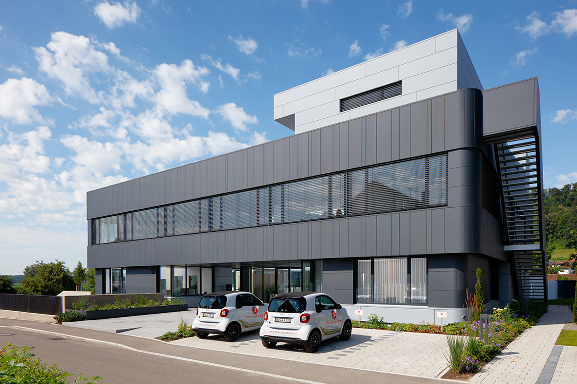 Modernes, mehrstöckiges Gebäude mit großen Fenstern und grauer Fassadenverkleidung. Vor dem Gebäude stehen zwei kleine Autos auf einem gepflasterten Parkplatz. Im Hintergrund sind Bäume und ein blauer Himmel mit einigen Wolken sichtbar.