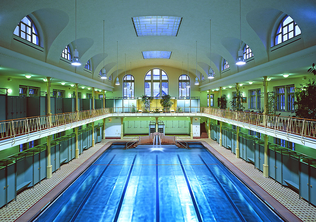 Ein großer, eleganter Innenraum eines Schwimmbads mit einem blauen Schwimmbecken in der Mitte. Die Wände sind mit Fenstern und Pflanzen geschmückt, und große, gewölbte Decken sorgen für eine helle, offene Atmosphäre.