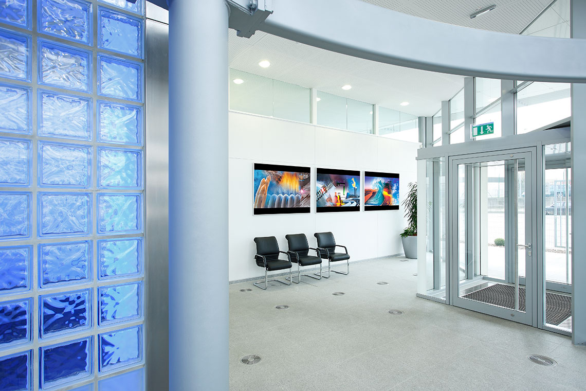 Ruhiger Empfangsbereich mit Glaswand und mehreren Sitzplätzen. An der Wand hängen drei Bilder mit lebhaften Farben. Die Eingangstür ist aus Glas, und in der Ecke steht eine Pflanze.