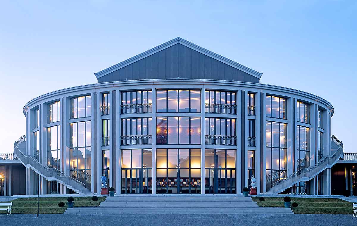 Eine runde, moderne Gebäudeansicht mit großen Glasfenstern. Zwei Treppen führen zum Haupteingang. Der Himmel ist hellblau und der Garten vor dem Gebäude ist ordentlich gestaltet. Die Architektur kombiniert traditionelle und zeitgenössische Elemente.