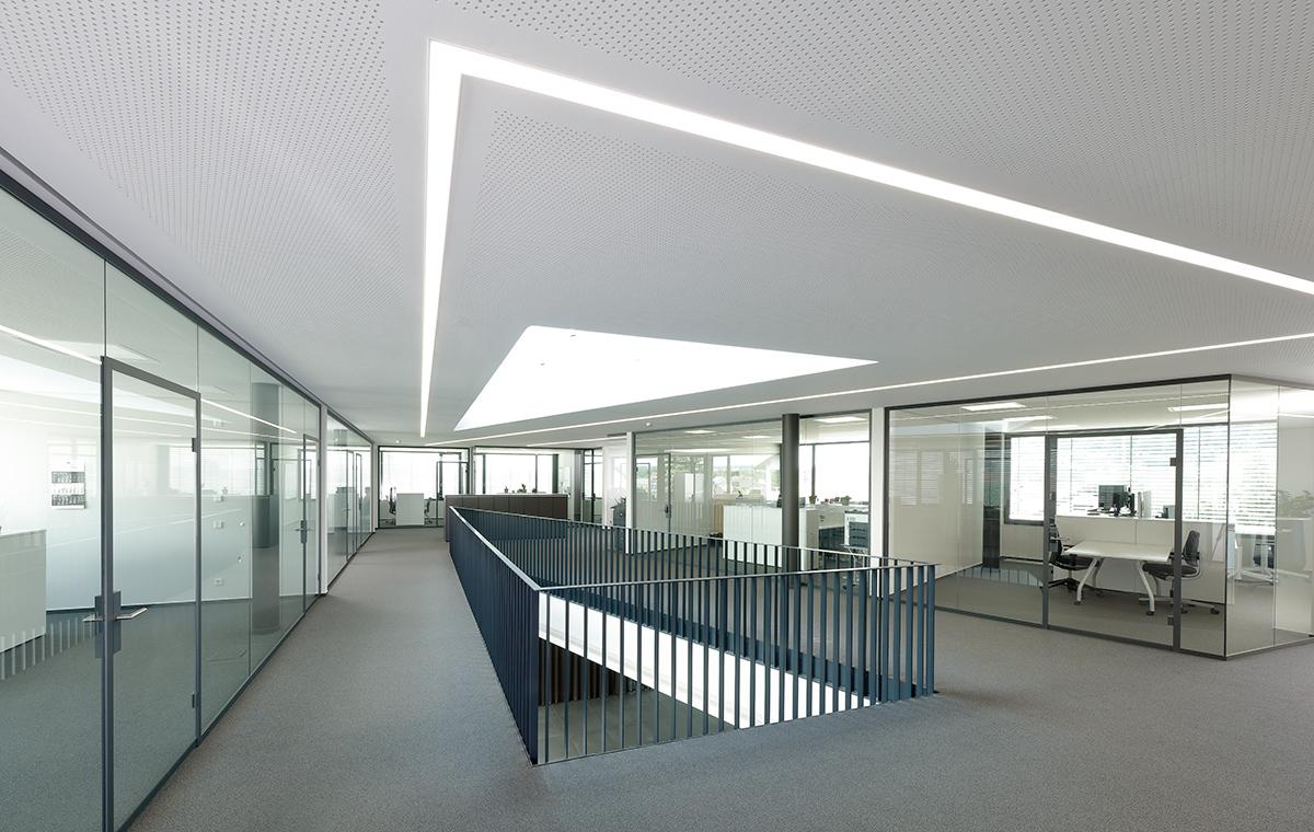 Moderne Bürolandschaft mit Glaswänden und einem offenen Bereich. Helle, minimalistische Gestaltung mit natürlichem Licht, das durch ein großes Dachfenster strömt. Ein erhöhter Gang führt zwischen den Büroräumen. Der Boden ist mit grauem Teppich ausgelegt.