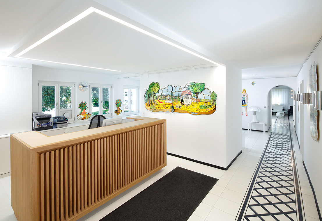 Eine modern gestaltete Empfangshalle mit einem hölzernen Empfangstresen. An der Wand hängt ein farbenfrohes Wandbild einer Landschaft. Der Boden ist hell gefliest und im Hintergrund sind weitere Räume sichtbar.