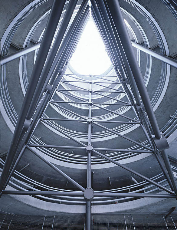 Ein Blick nach oben in einen modernen, zylindrischen Raum mit sichtbaren Stahlträgern. Das Licht strömt von oben durch eine kreisförmige Öffnung, während die Wände aus Beton und Stahl konstruiert sind. Die Struktur vermittelt ein Gefühl von Höhe und Raum.