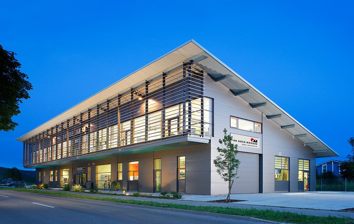 Moderner, zweigeschossiger Gebäude in der Dämmerung. Große Fenster und eine klare, rechteckige Struktur. Vor dem Gebäude ist ein kleiner Baum sowie eine asphaltierte Straße zu sehen. Die Fassade hat vertikale Holzleisten und eine klare Beschriftung am Eingang.