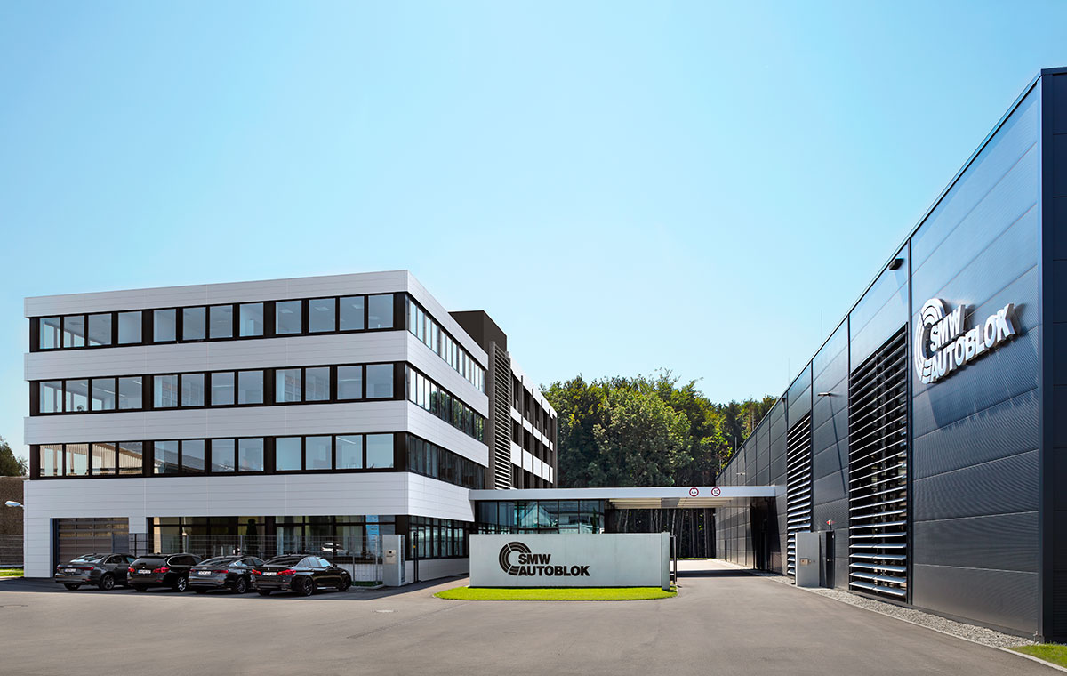 Moderne Unternehmensgebäude mit zwei Abschnitten, einem mehrstöckigen weißen Gebäude und einem schwarzen, modernen Anbau. Im Vordergrund steht ein Schild mit dem Firmenlogo. Vor dem Gebäude parken mehrere Autos. Der Himmel ist klar und blau.
