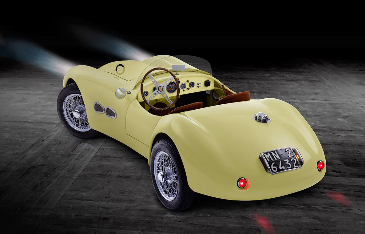 Ein klassisches, gelbes Sportauto mit offenem Design und einer hölzernen Innenausstattung. Das Fahrzeug hat Drahtspeichenräder und eine Motorhaube mit einem runden Emblem. Das Nummernschild ist gut sichtbar. Der Hintergrund ist dunkel, was das Auto hervorhebt.