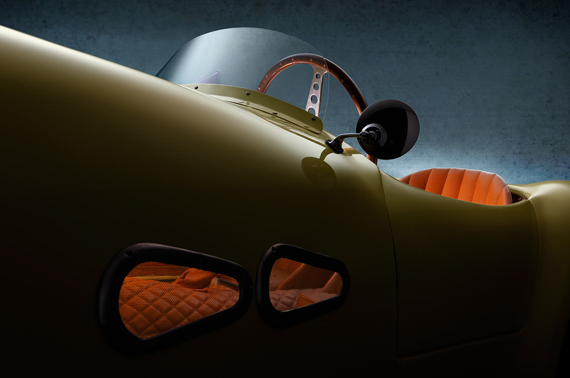 Nahaufnahme eines klassischen Autos mit einer geschwungenen Form. Die Karosserie ist in einem gelben Farbton, und die Sitze sind in einem auffälligen Orangeton gehalten. Der Innenraum zeigt ein modernes Lenkrad und eine Windschutzscheibe, während die Hintergrundfarbe dunkel und strukturiert ist.