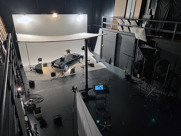 Ein Blick auf ein Fotostudio mit einem schwarzen Auto auf einem weißen Hintergrund. Ein Fotograf arbeitet am Auto, während mehrere Scheinwerfer und Kameras aufgestellt sind. Der Raum ist dunkel, und es sind verschiedene Studioeinrichtungen sichtbar.