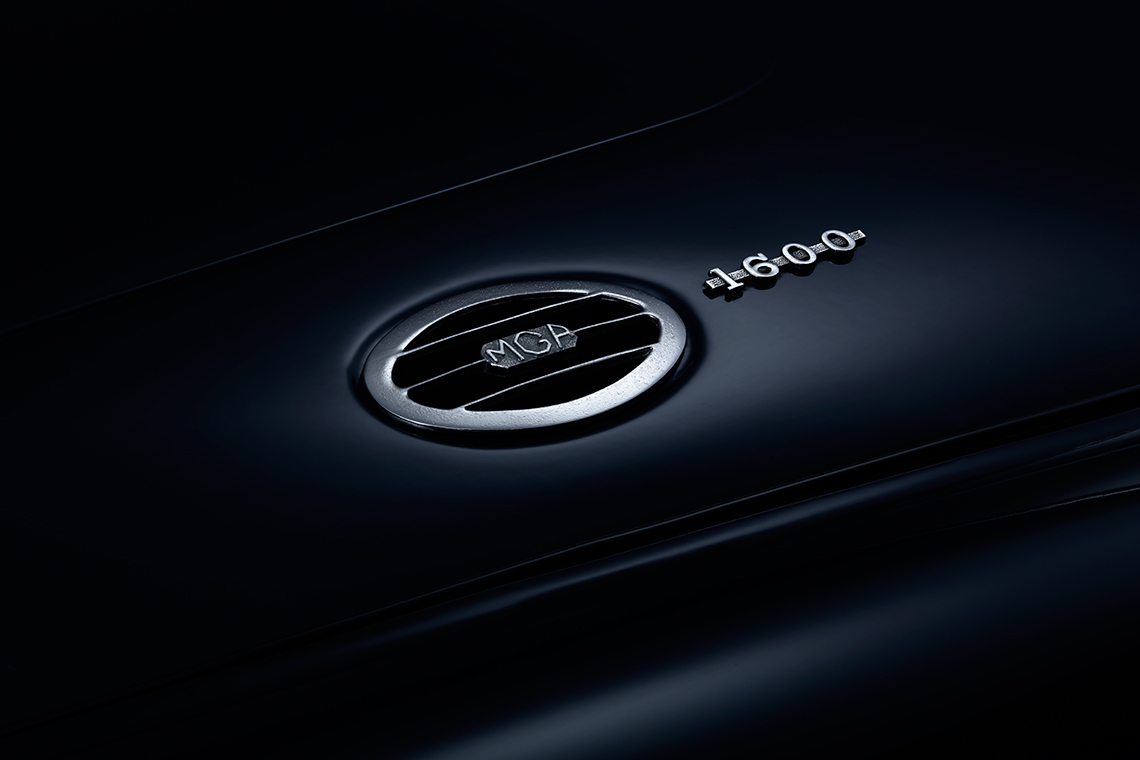 Ein schimmerndes Logo und die Modellbezeichnung "MGA 1600" auf einer dunklen Fahrzeugoberfläche. Das Logo hat eine runde Form mit horizontalen Streifen und ist in silberner Farbe gehalten.