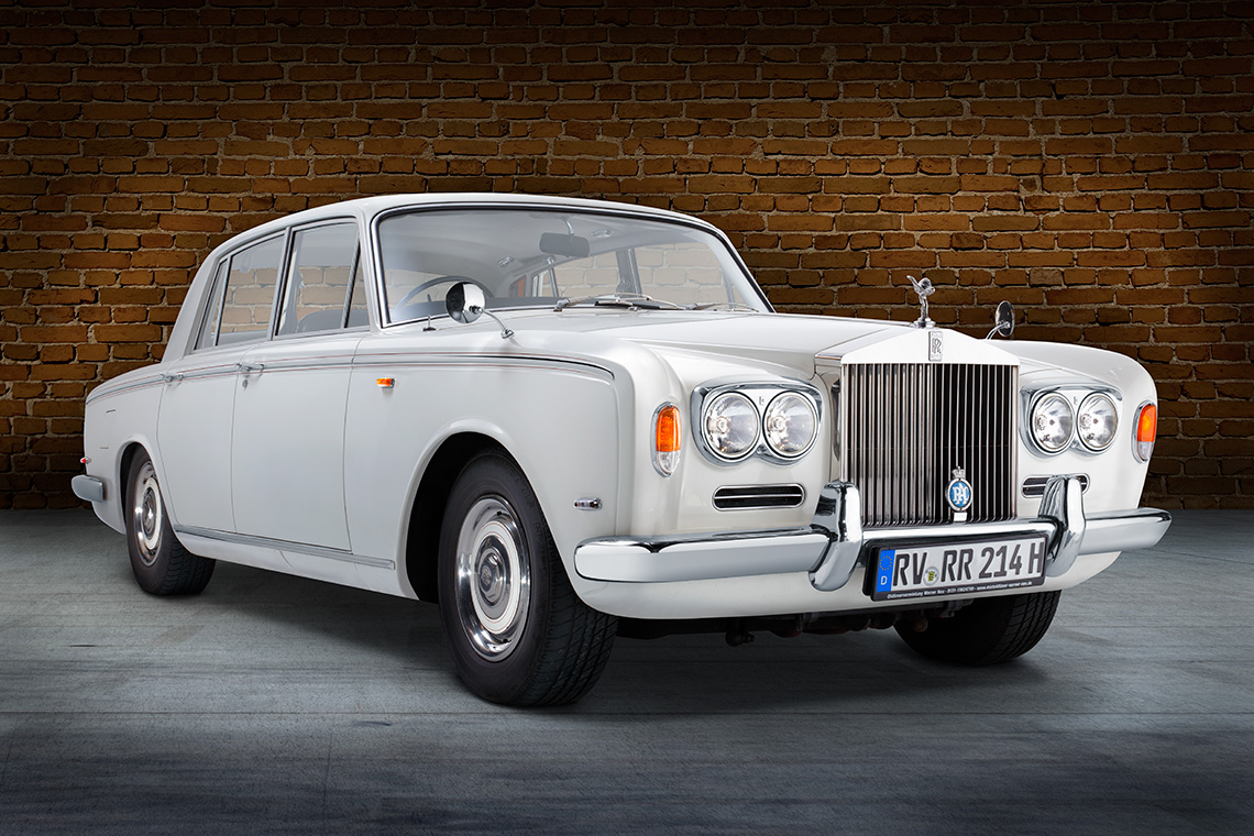 Ein klassisches weißes Auto, ein Rolls-Royce, steht vor einer Backsteinmauer. Die Front weist markante Scheinwerfer und das bekannte Kühleremblem auf. Das Fahrzeug hat eine elegante und luxuriöse Erscheinung.