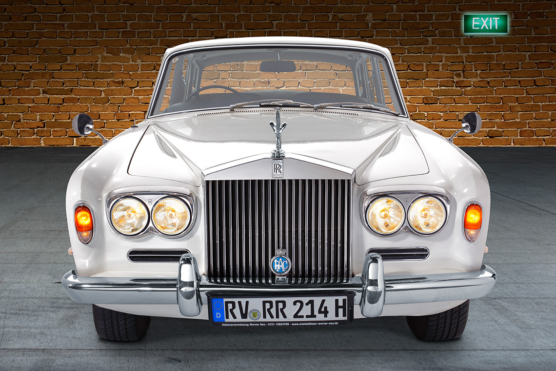 Ein klassischer, weißer Rolls-Royce mit markanter Kühlergrill und runden Scheinwerfern. Das Fahrzeug steht vor einer Backsteinwand. Ein grünes Ausstiegsschild ist im Hintergrund sichtbar.