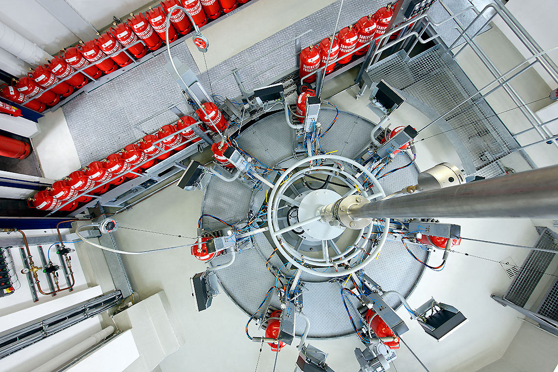 Eine Vogelperspektive auf eine kreisförmige Anordnung von technischen Geräten und Rohren. Um den zentralen Bereich sind mehrere rote Gasflaschen auf einer geraden Plattform angeordnet. Der Boden ist grau und die Umgebung wirkt industriell.