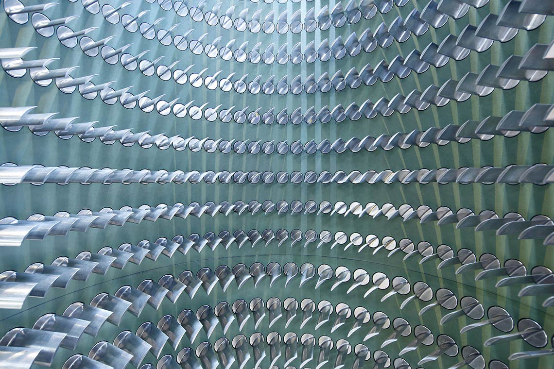 Ein Muster von vielen silbernen Rohren, die kreisförmig angeordnet sind. Die Rohre sind teilweise spiegelnd und reflektieren das Licht, was einen dreidimensionalen Effekt erzeugt. Der Hintergrund ist in einem sanften Grünton gehalten.