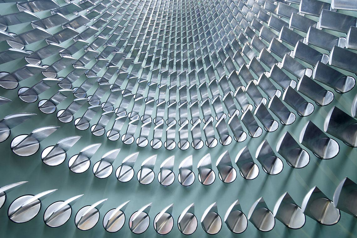 Eine abstrakte Struktur mit sich spiralförmig anordnenden, glänzenden Metallteilen. Die Elemente sind halbrund und reflektieren das Licht, was einen dynamischen Eindruck erzeugt. Der Blickwinkel ist von unten nach oben gerichtet, wodurch die Tiefe der Form betont wird.