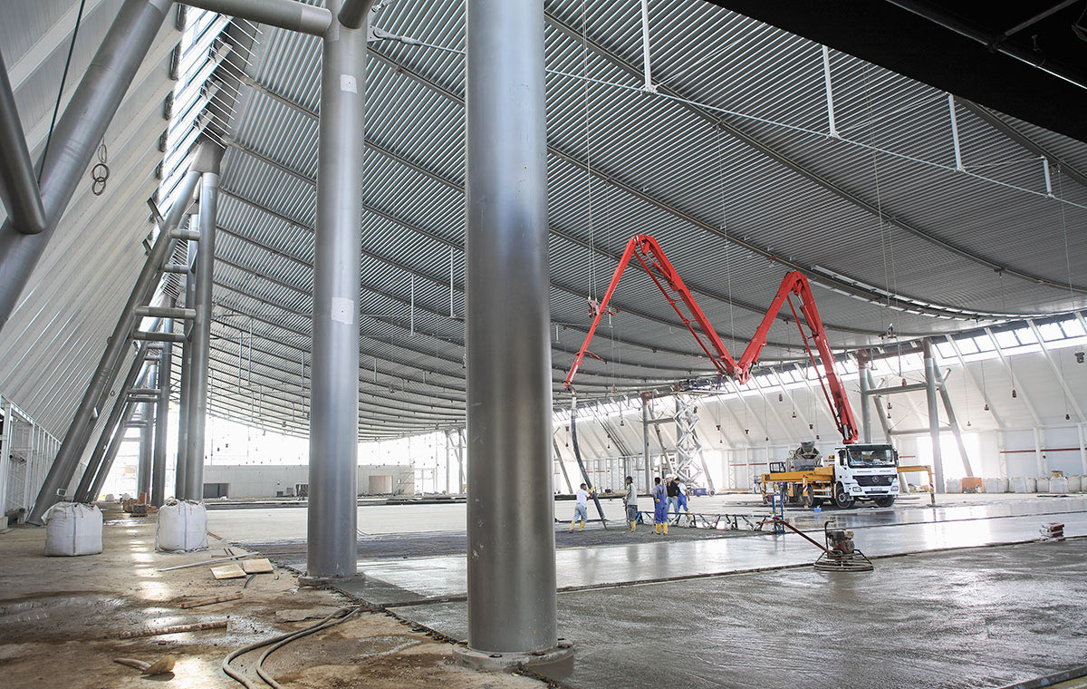 Ein großer, leerer Innenraum mit hohen, metallic schimmernden Wänden und einem gewölbten Dach. In der Mitte befindet sich ein Betonmischer mit einem langen Schlauch, während Arbeiter auf dem Boden beschäftigt sind. Stützen aus Edelstahl sind sichtbar.