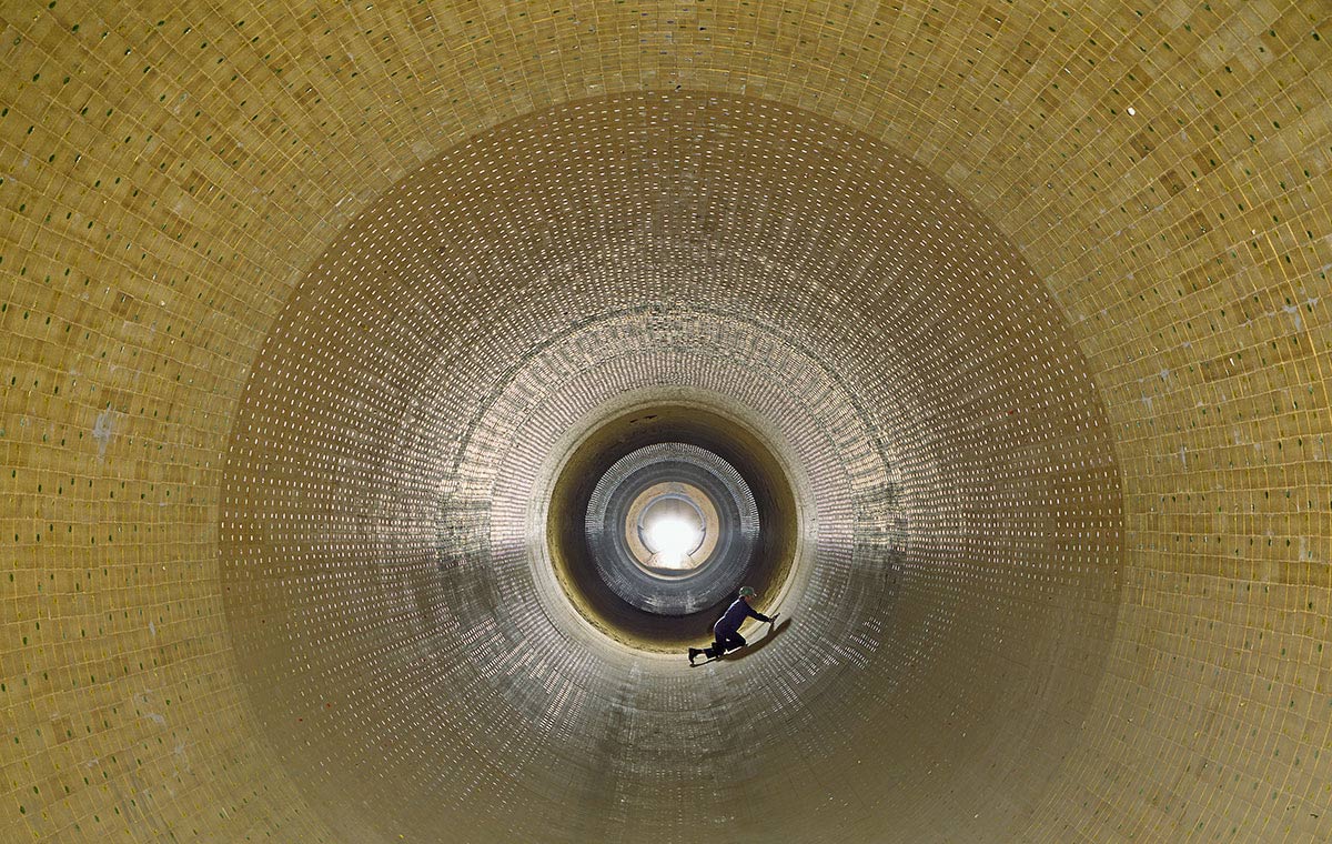 Eine perspektivische Ansicht des Innenraums eines großen, zylindrischen Behälters aus Steinen oder Platten. In der Mitte ist eine Lichtquelle sichtbar, während eine Person am Boden arbeitet. Die Wände sind rhythmisch strukturiert und reflektieren das künstliche Licht.