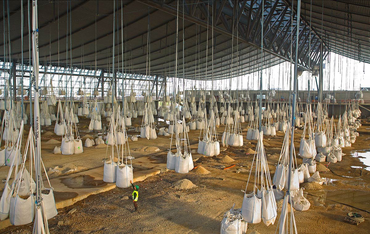 Ein großer Lagerraum mit vielen hängenden Säcken, die mit Material gefüllt sind. Ein Arbeiter in gelber Sicherheitskleidung steht am Boden zwischen den Säcken. Die Wände des Raumes sind aus Metall und der Boden ist uneben.