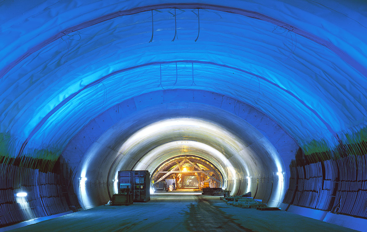 Ein beleuchteter Tunnel mit blauen Wänden und einem gewölbten Dach. Im Hintergrund sind einige Baugeräte und Personen zu sehen. Der Boden ist eben und der Tunnel scheint gut strukturiert zu sein.