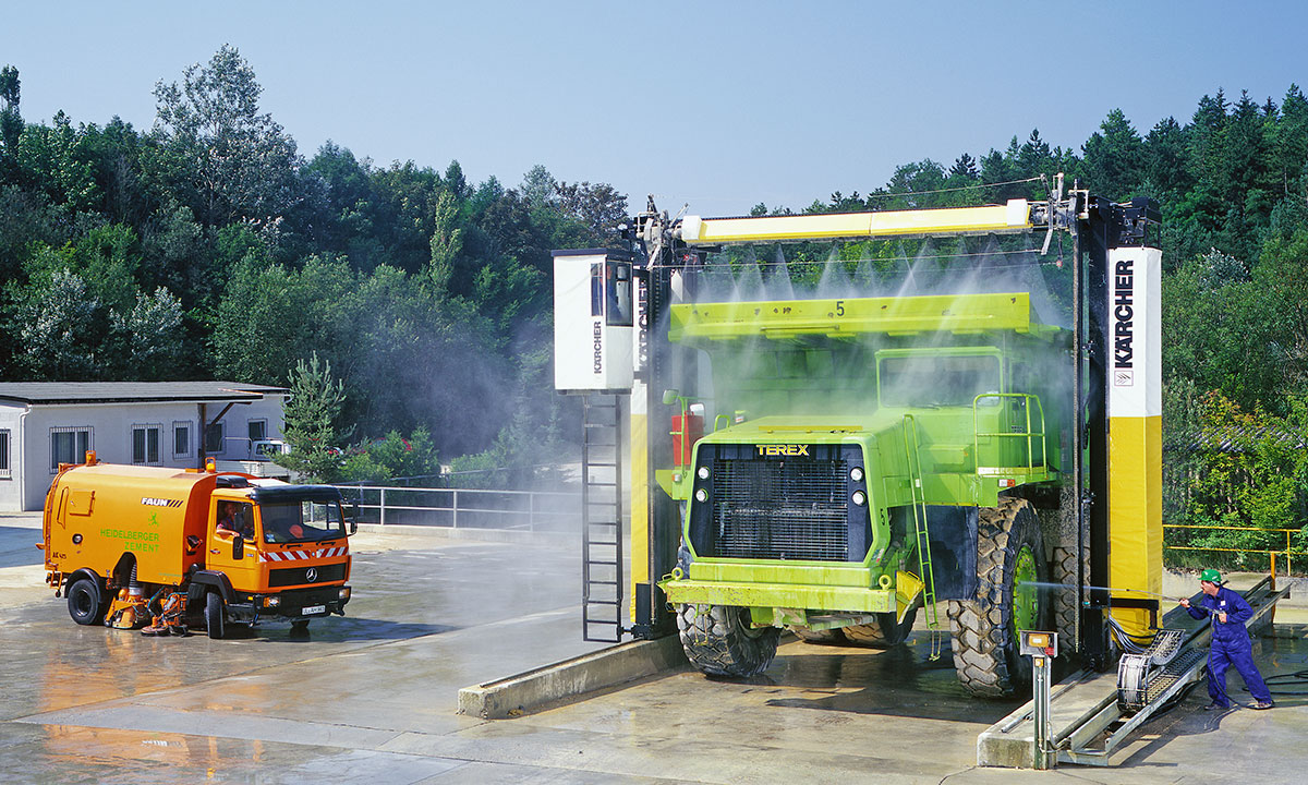 Ein grüner Großtraktor wird von einer automatischen Waschanlage gereinigt. Neben der Waschanlage steht ein orangefarbener Reinigungswagen. Wasser spritzt überall, während ein Arbeiter in blauer Arbeitskleidung zuschaut. Im Hintergrund sind Bäume und eine Gebäudeansicht sichtbar.