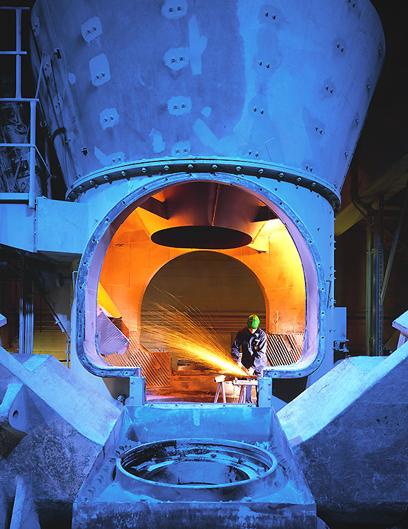 Ein Arbeitsbereich in einer Industrieanlage, in dem ein Mitarbeiter mit Schutzkleidung an einem Maschineninneren arbeitet. Aus dem Inneren der Maschine strömt ein helles, orangefarbenes Licht, während Funken sprühen. Die Umgebung ist stark beleuchtet und metallisch geprägt.