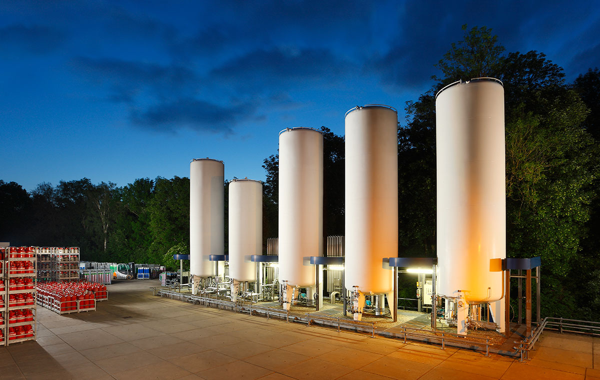 Fünf große, weiße Zylinderspeicher stehen in einer Außenanlage, umgeben von Bäumen. Im Hintergrund sind einige Paletten oder Kästen zu sehen. Die Szene ist bei Dämmerlicht aufgenommen, wobei die Speicher leicht beleuchtet sind.