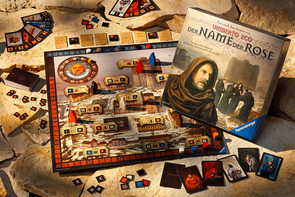 Ein Brettspiel mit dem Titel "Der Name der Rose" liegt auf einem felsigen Untergrund. Der Spielplan zeigt verschiedene Orte und Karten, während die Spielbox und Spielkarten im Hintergrund zu sehen sind.