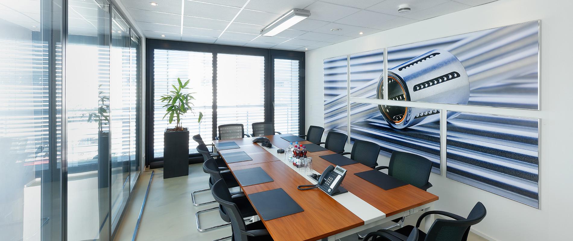 Ein modern gestalteter Konferenzraum mit einem langen Tisch, umgeben von Stühlen. An der Wand hängt ein großes Bild eines Flugzeugmotors. Große Fenster lassen viel Tageslicht herein, und in einer Ecke steht eine Pflanze.
