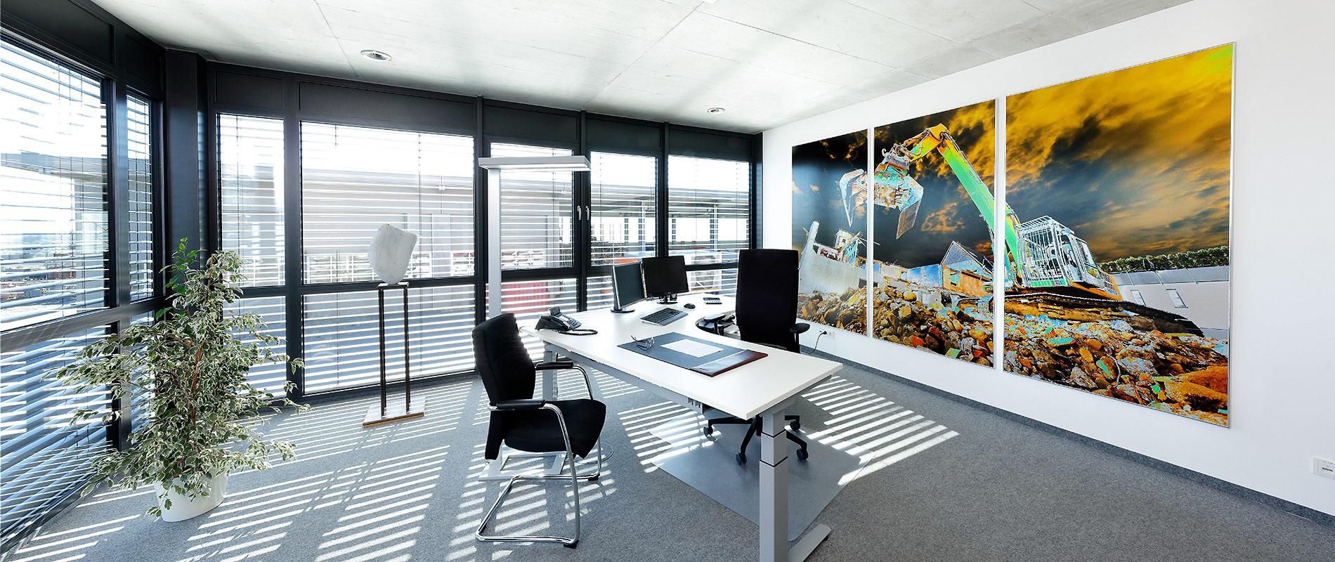 Ein modernes Büro mit großen Fenstern, die viel Tageslicht hereinlassen. Der Raum ist mit einem Schreibtisch, Stühlen und einer Pflanze eingerichtet. An der Wand hängt ein großes, buntes Bild, das eine Baustelle zeigt.