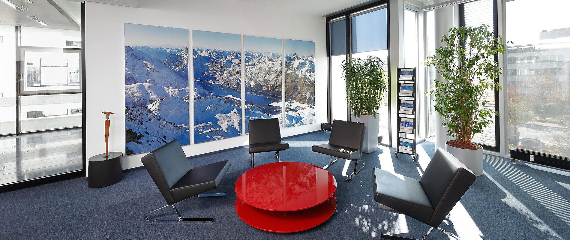 Moderne Wartebereich mit einem runden, roten Tisch in der Mitte und schwarzen Sesseln um ihn herum. Große Fenster lassen viel Licht herein, und an der Wand hängt ein großes Bild von einer Berglandschaft. Eine Pflanze steht ebenfalls im Raum.