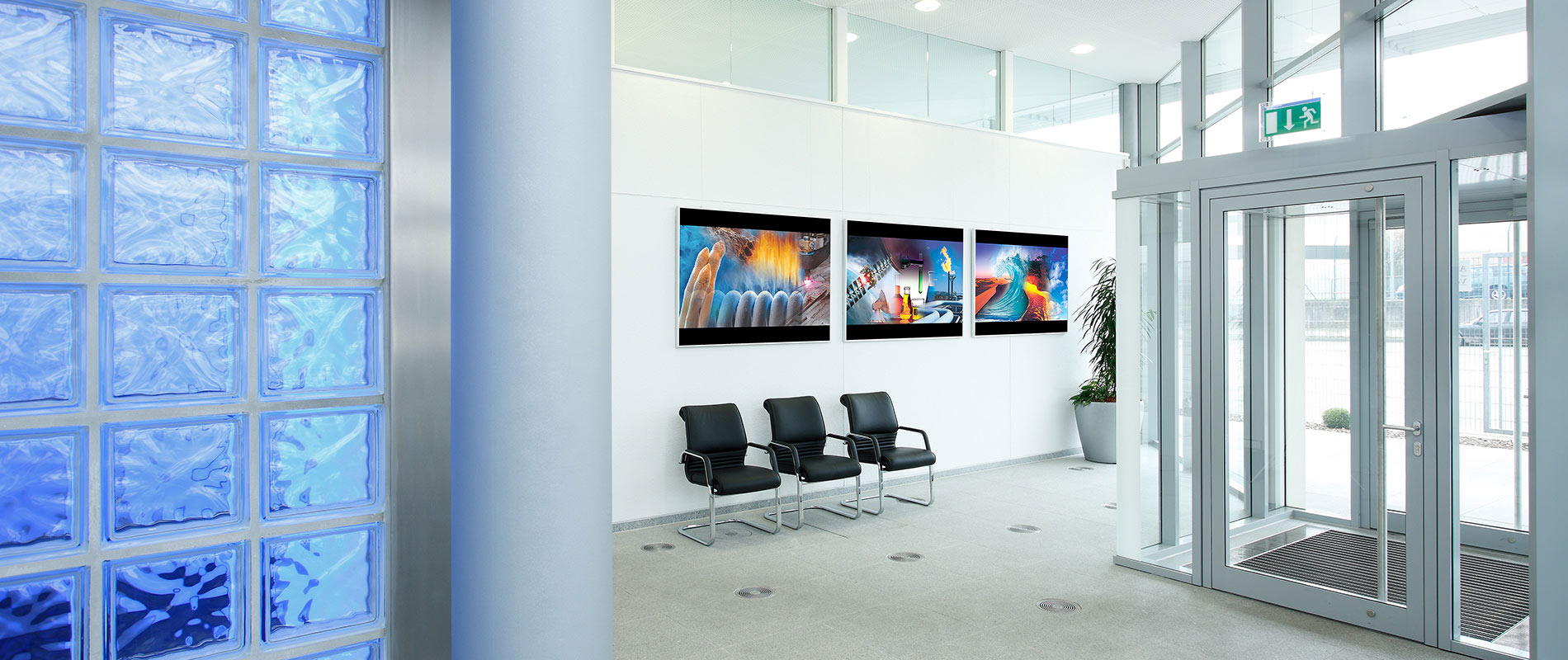 Ein moderner Empfangsbereich mit großen Fenstern und hellen Wänden. Auf einer Wand hängen drei Bilder, während auf dem Boden zwei schwarze Stühle stehen. Links ist eine Glassteinwand sichtbar, die durch das Licht ein attraktives Muster erzeugt.