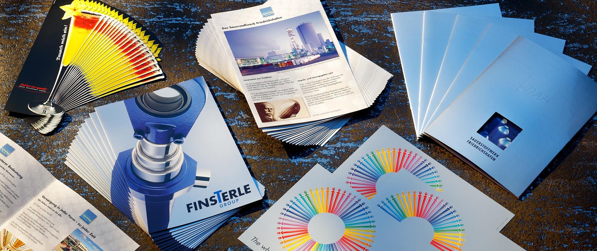 Eine Sammlung von Broschüren und Farbmustern auf einem Holzuntergrund. Die Werke zeigen verschiedene Designs, darunter Produkte und Informationen über das Unternehmen. Farbenfrohe Fächer und informative Dokumente sind sichtbar.