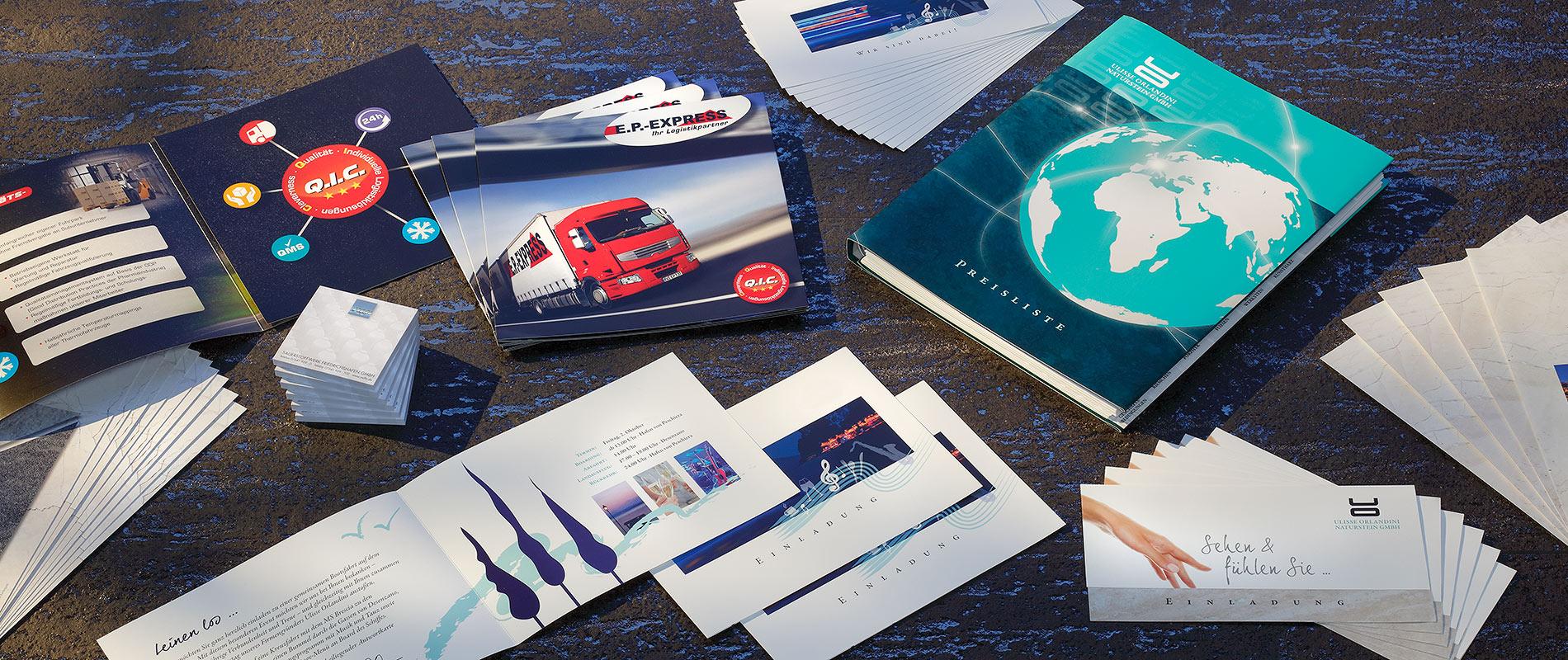 Eine Vielzahl von gedruckten Materialien liegt auf einem Tisch. Dazu gehören Broschüren, Flyer und Visitenkarten mit verschiedenen Designs, darunter Bilder von Lkw und einer Weltkarte. Die Farben sind überwiegend blau, rot und weiß.