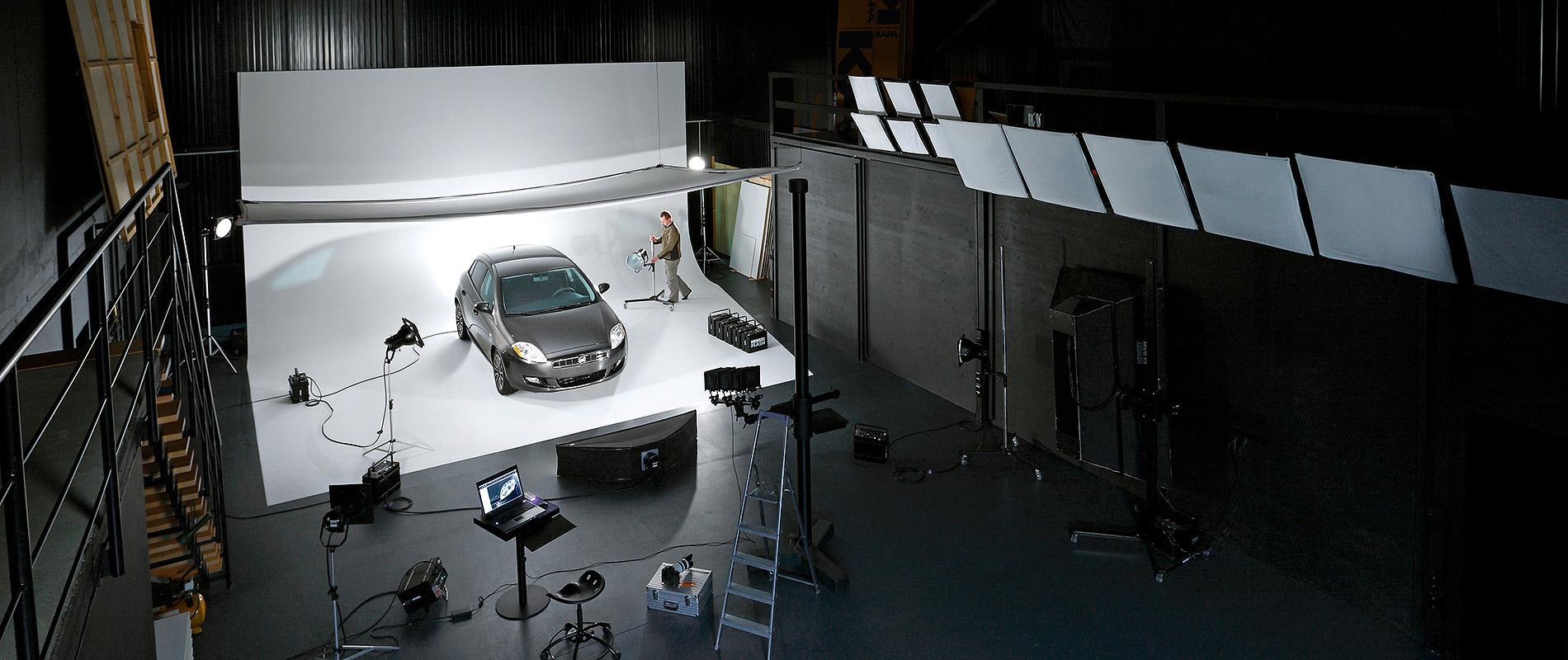 Ein Auto steht auf einem beleuchteten Fotostudio-Hintergrund. Rund um das Fahrzeug sind verschiedene Lichtquellen und Kamerastative angeordnet. Eine Person arbeitet im Hintergrund, um die Aufnahme vorzubereiten.