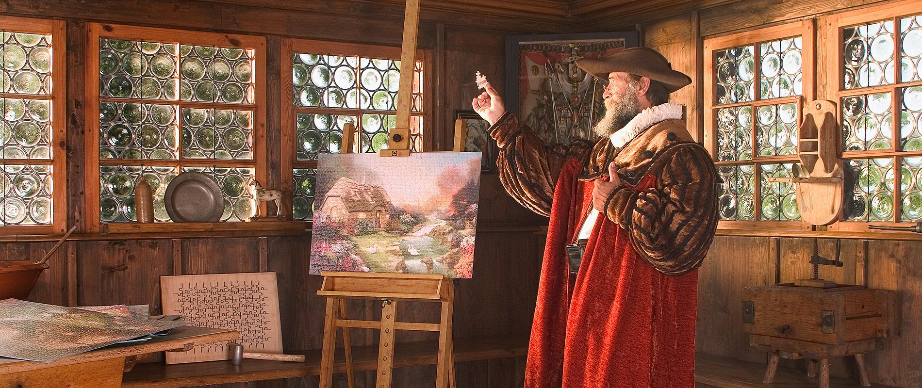Ein Mann in historischer Kleidung steht in einem Holzzimmer und betrachtet ein Gemälde auf einer Staffelei. Die Wände sind mit Fenstern und Kunstwerken dekoriert, während verschiedene Malutensilien auf einem Tisch liegen. Das Licht strömt durch die Fenster und schafft eine warme Atmosphäre.