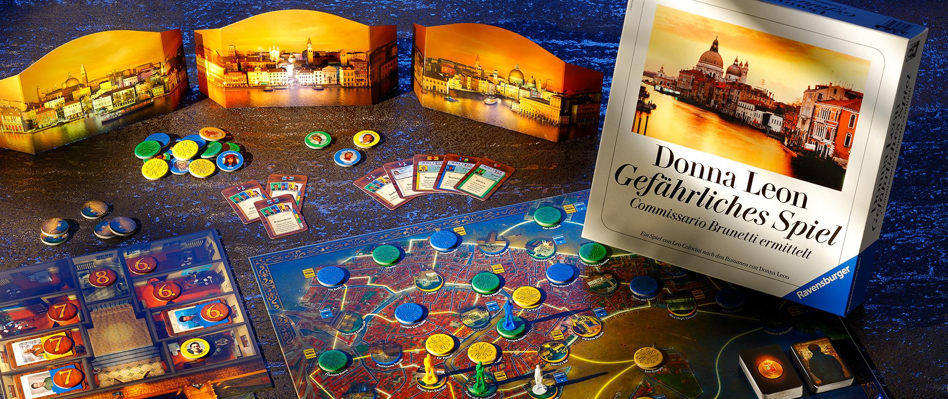 Ein Brettspiel liegt auf einem Tisch, umgeben von Spielkarten, Spielsteinen und farbigen Chips. Im Hintergrund stehen zwei aufgestellte Spielwände mit Bildern von Städten. Ein Buch mit dem Titel "Donna Leon Gefährliches Spiel" ist prominent platziert.