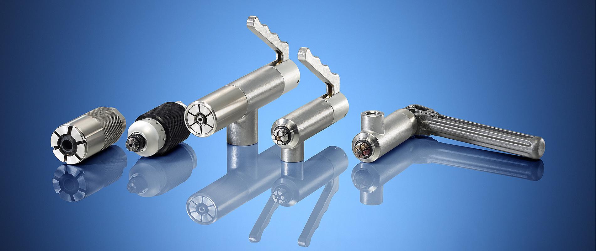 Eine Gruppe von vier metallischen Werkzeugen auf einem blauen Hintergrund. Die Werkzeuge haben unterschiedliche Formen und Größen, darunter Zylinder und eine Griffkonstruktion. Sie sind alle glänzend und reflektieren das Licht.