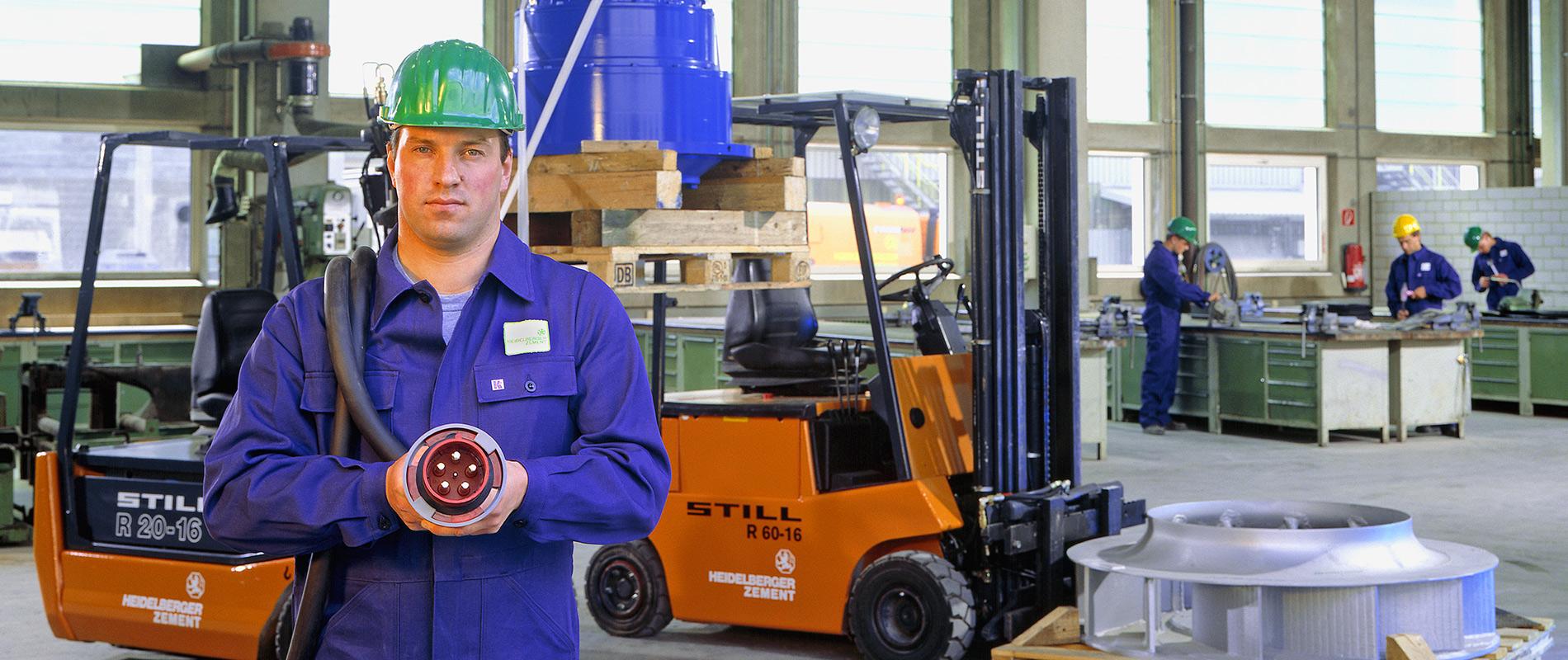 Ein Mann in blauer Arbeitskleidung und einem grünen Helm steht in einer Lagerhalle. Er hält ein technisches Bauteil in der Hand. Im Hintergrund sind Gabelstapler und Arbeiter an Werkbänken zu sehen. Die Umgebung ist hell und industriell gestaltet.