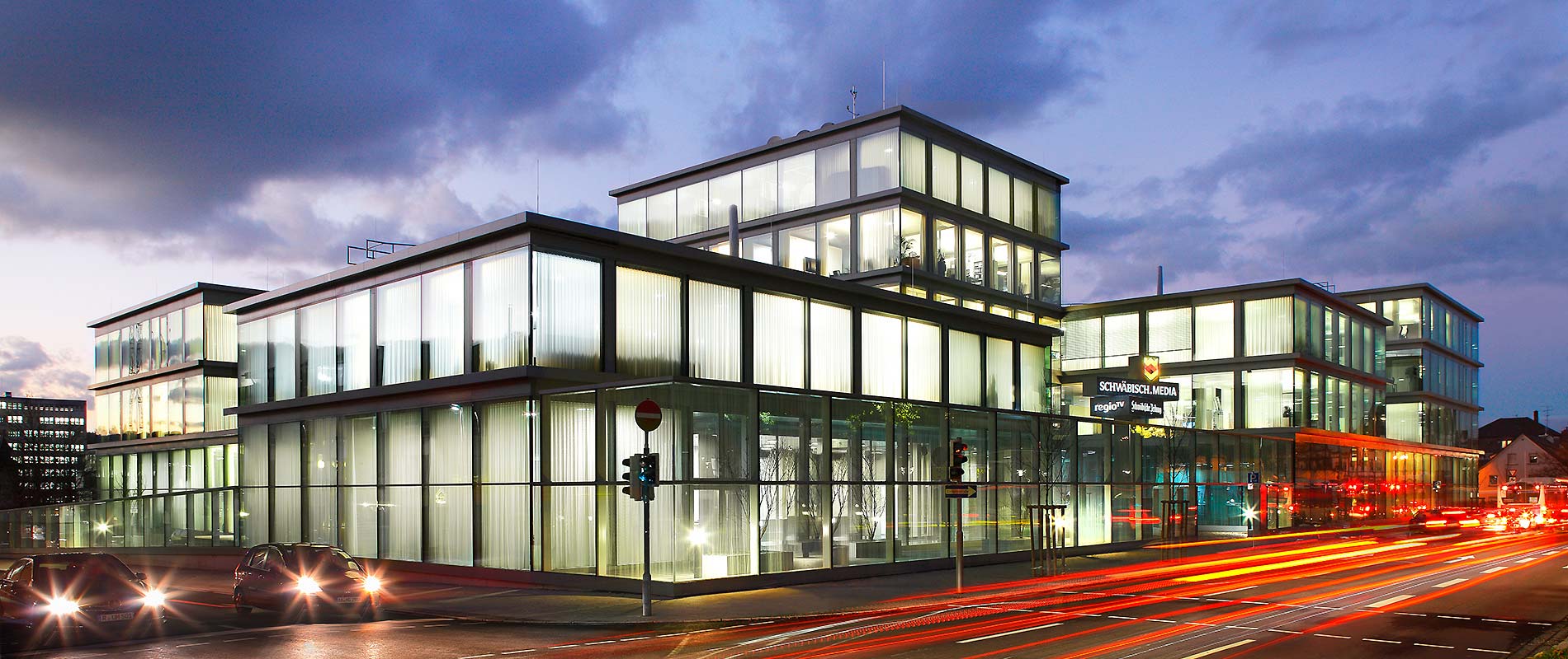 Moderne Bürogebäude mit Glasfassade bei Dämmerung. Die Architektur ist klar und geometrisch, umgeben von Straßen mit leuchtenden Fahrzeuglichtern. Der Himmel ist bewölkt und düster, was die beleuchteten Fenster hervorhebt.