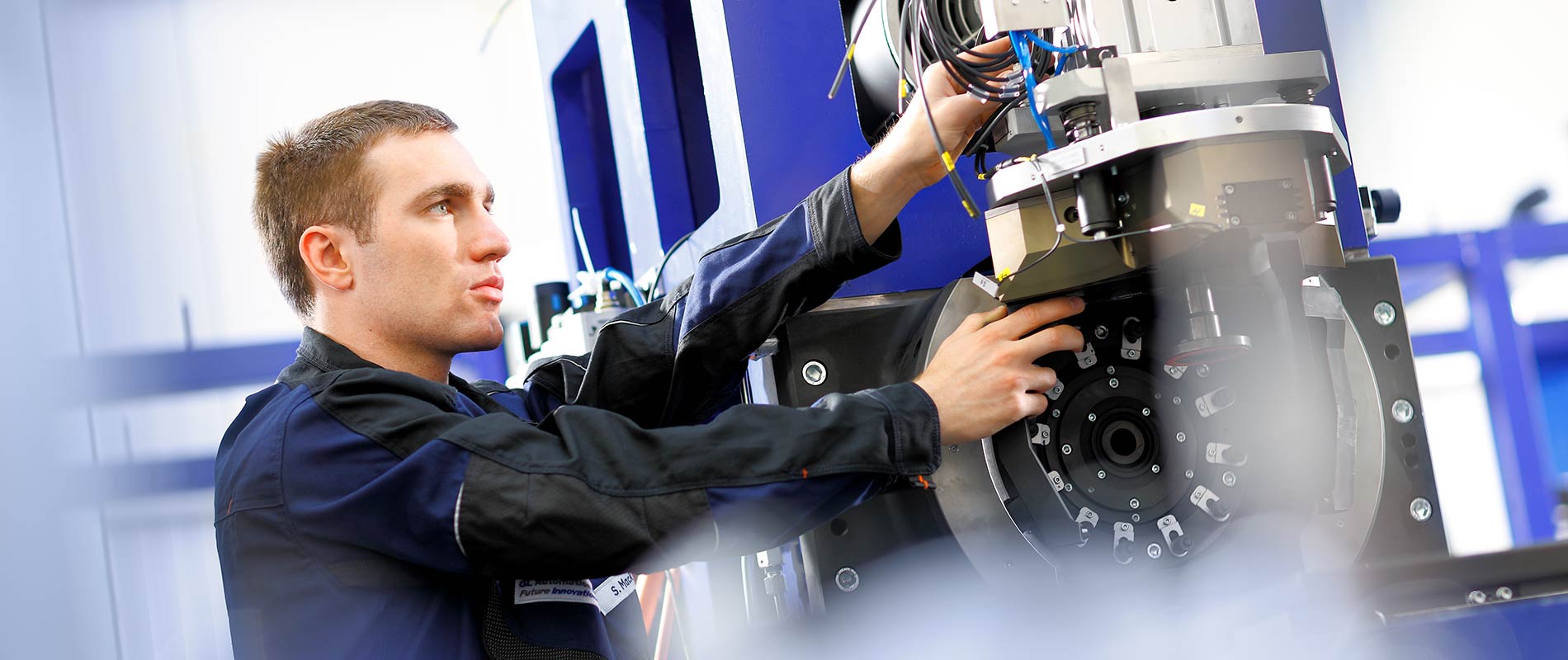 Ein Techniker arbeitet an einer Maschine, während er Kabel und Teile überprüft. Er trägt eine Arbeitsjacke und konzentriert sich auf die Installation oder Wartung der technischen Komponenten. Der Hintergrund zeigt industrielle Ausrüstung und Maschinen.
