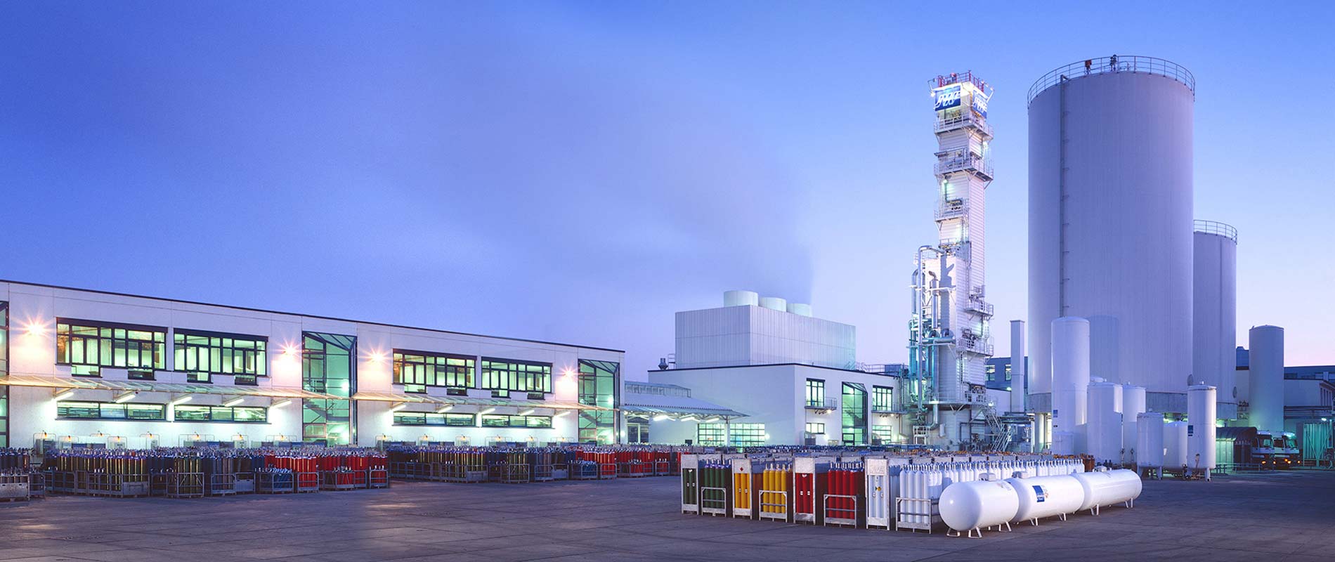 Industriegelände bei Dämmerung mit großen Lagertanks, Produktionsgebäuden und Gaszylindern auf dem Boden. Die Gebäude sind modern und gut beleuchtet.