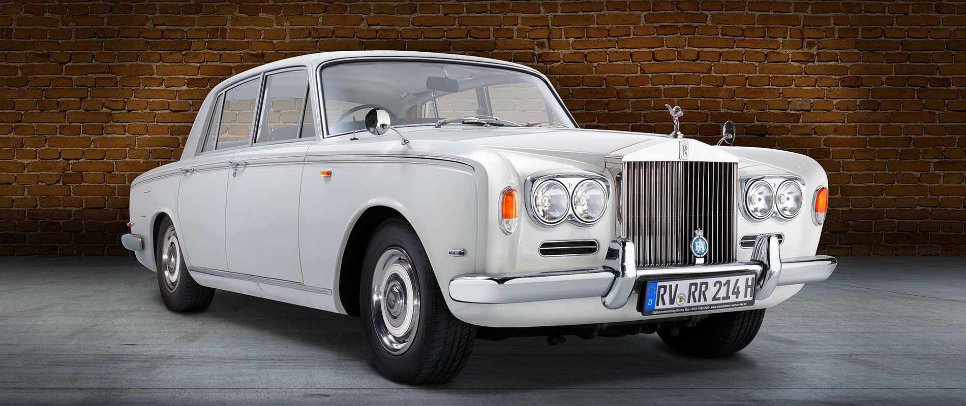 Ein eleganter, klassischer Rolls-Royce in Weiß steht vor einer Backsteinmauer. Der Luxuswagen hat vier Türen, runde Scheinwerfer und ein markantes Kühlergrill mit dem Rolls-Royce-Emblem. Der Hintergrund besteht aus einer stilvollen grauen Fläche.