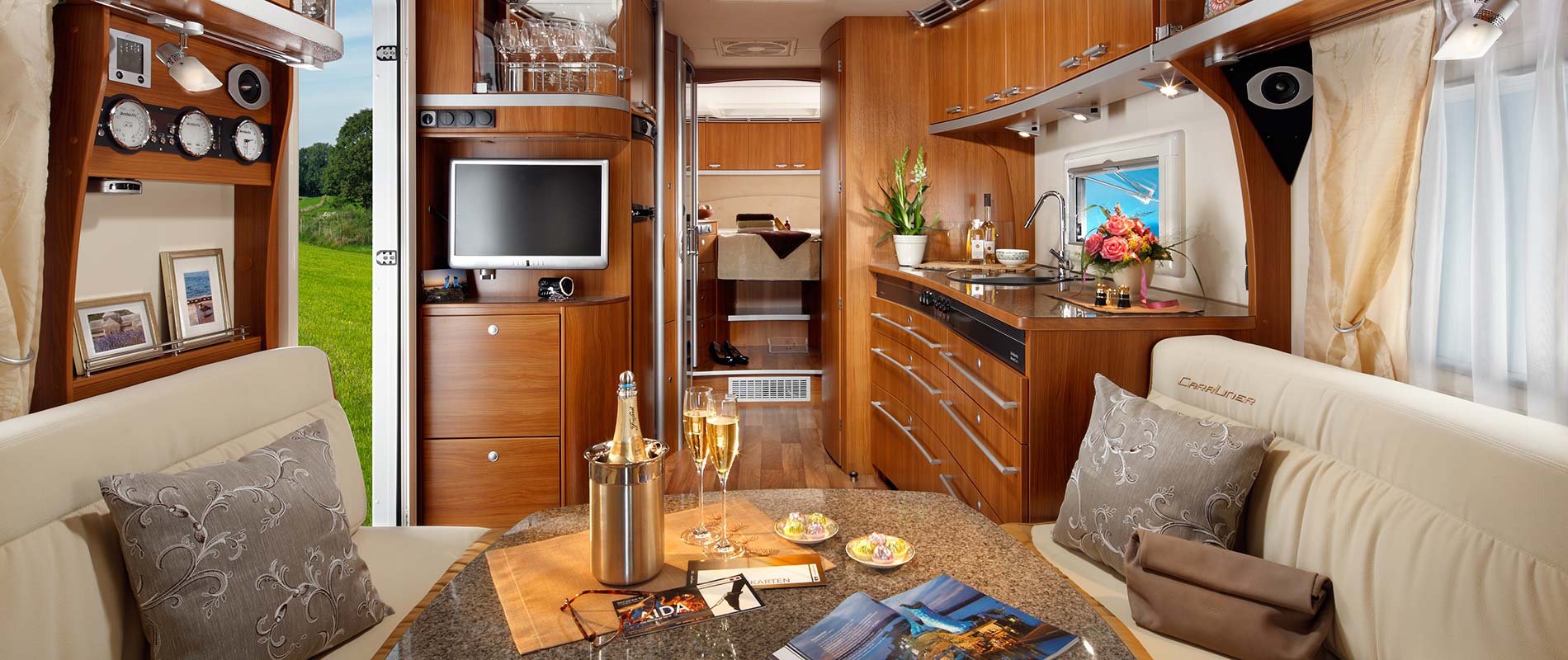 Eine modern gestaltete Innenansicht eines Wohnmobils mit holzverkleideten Wänden. Die Küche ist gut ausgestattet mit Geräten, während ein gemütlicher Sitzbereich mit einem Tisch und Dekorationen zu sehen ist. Fenster bieten einen Blick ins Grüne.